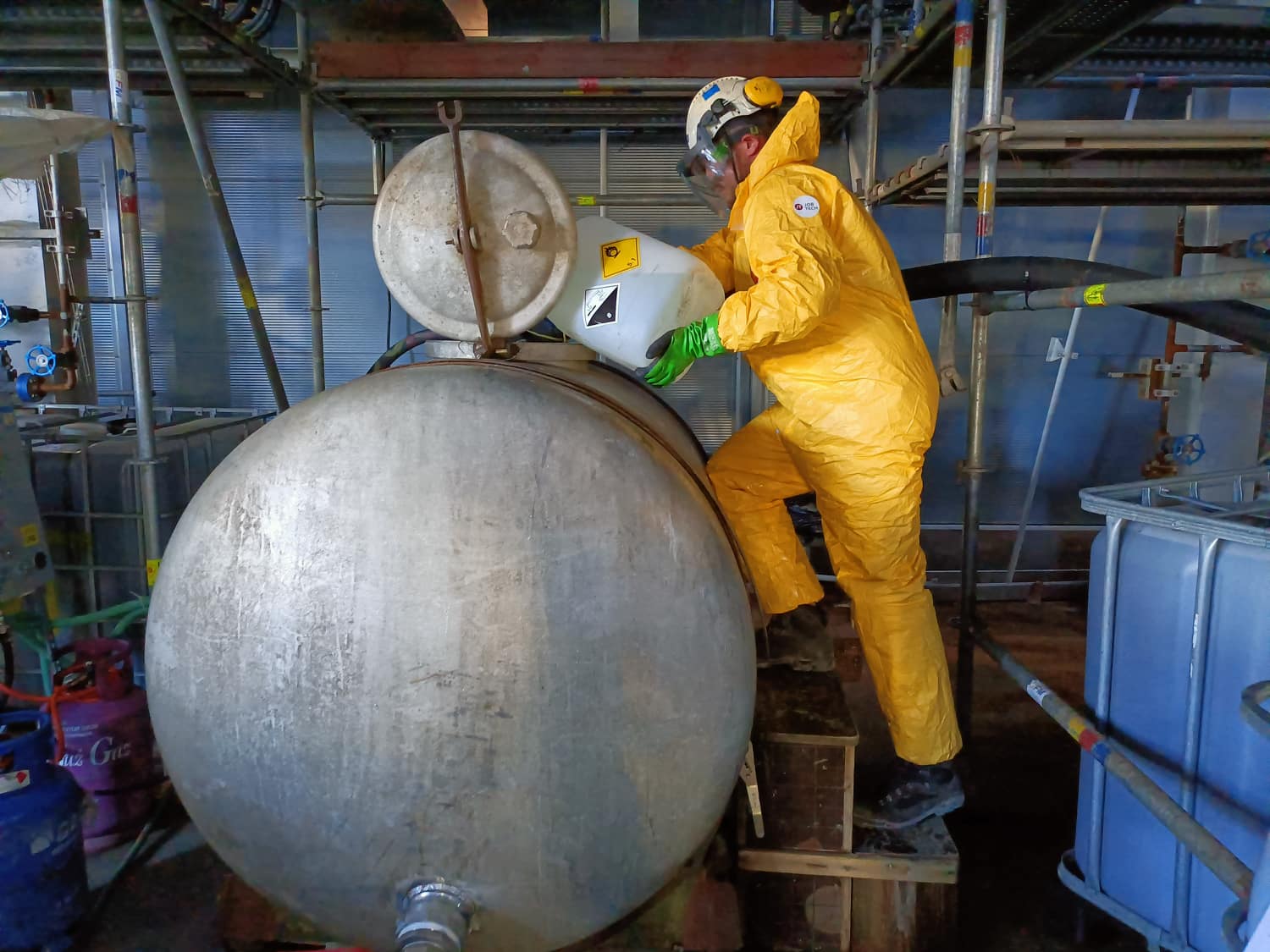 Technik JobTech w ubraniu chemoodpornym dozujący chemikalia do zbiornika podczas czyszczenia chemicznego rurociągu